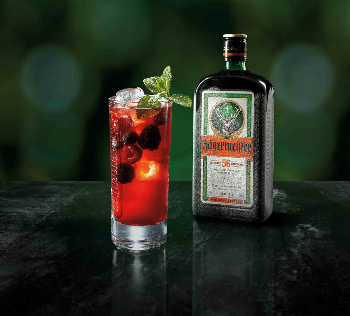 A bottle of Jägermeister stands next to a highball glass filled with a red cocktail, ice, berries, and mint, set on a dark reflective surface against a blurred green background.