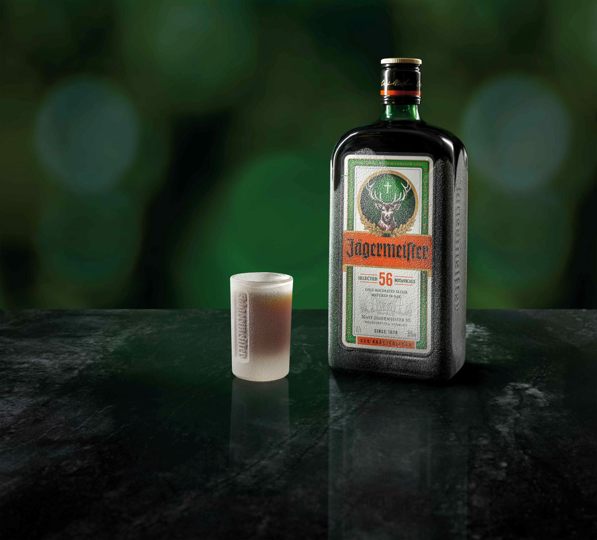 A bottle of Jägermeister stands on a dark reflective surface beside a small frosted shot glass, with a blurred green background.