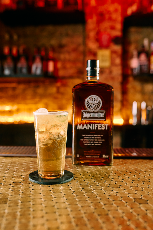 A bottle of Jagermeister Manifest and a glass of liquid with ice, set on a woven bar mat with a blurred bar background.