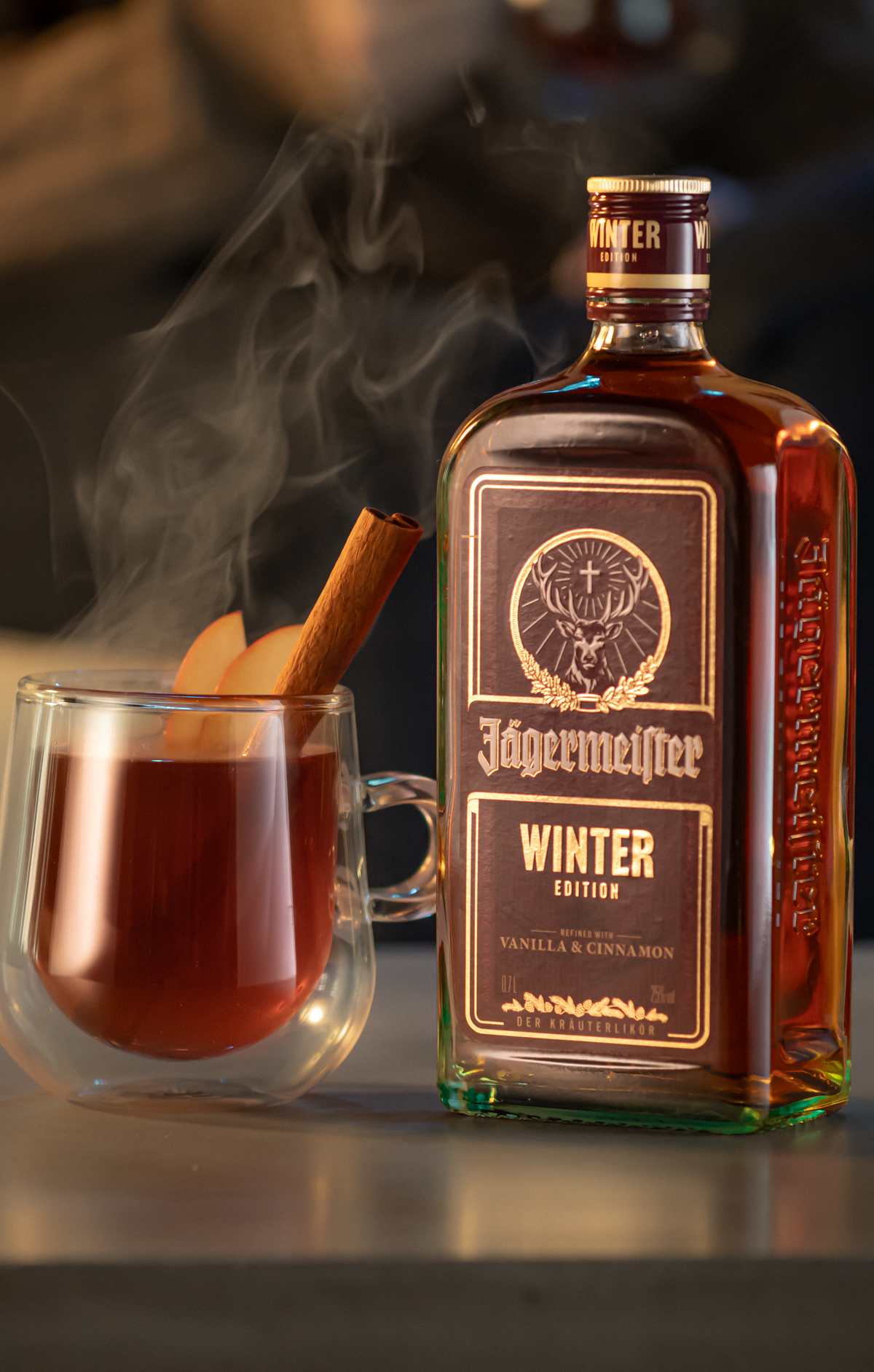 A bottle of Jägermeistler Winter Edition liquor stands beside a double-walled glass mug filled with a warm amber drink, garnished with a cinnamon stick and a slice of orange, with steam rising in a dimly lit bar setting.