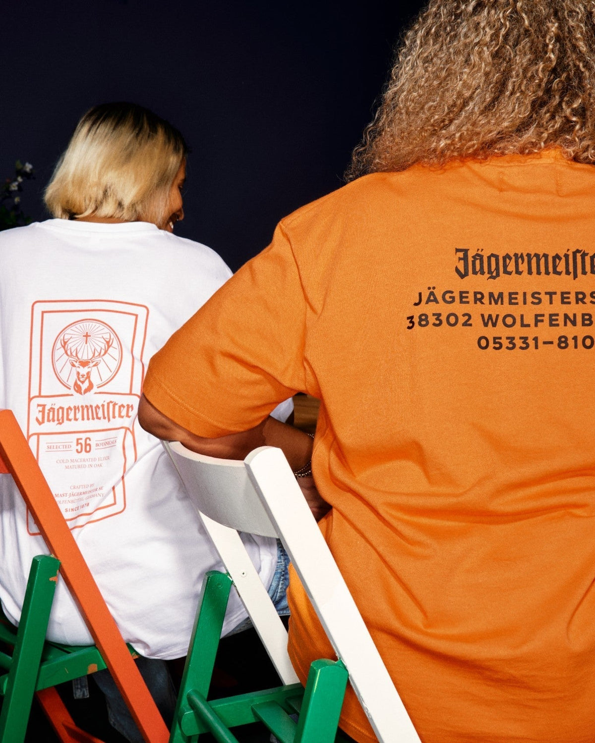 Due persone sedute su sedie verdi e bianche, riprese da dietro e con addosso magliette brandizzate Jägermeister. Una maglietta è bianca con stampa dell’etichetta arancione, e l’altra è arancione brillante con l’indirizzo della sede centrale Jägermeister in nero.