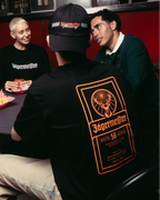 Quattro persone riunite attorno a un tavolo in un chiosco di street food, con il focus su una persona che indossa una T-shirt Jägermeister nera con una grande grafica arancione sul retro, pantaloni kaki e un berretto nero.