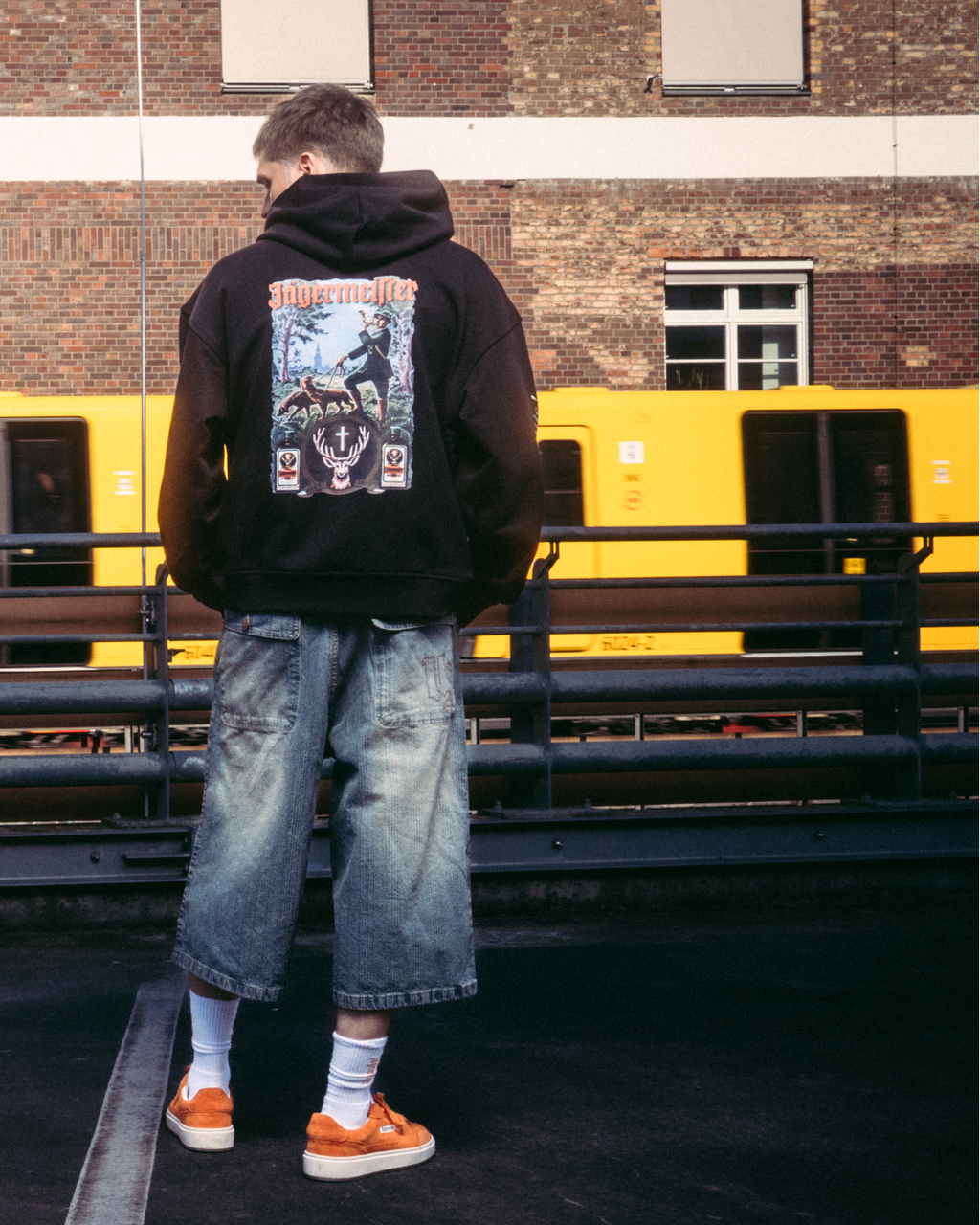 Una modella che indossa capi streetwear, tra cui una felpa con cappuccio nera con stampa grafica Jägermeister tradition sul retro, in piedi su una superficie asfaltata scura davanti a un edificio in mattoni e a un treno giallo.