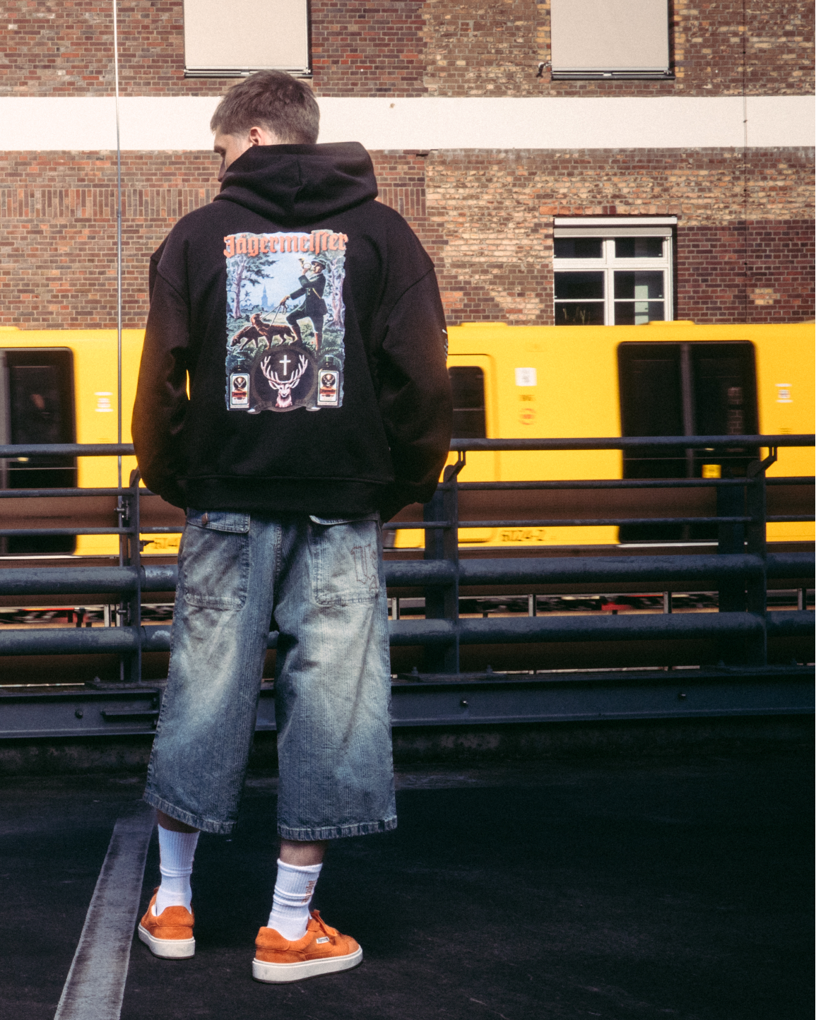 Una modella che indossa capi streetwear, tra cui una felpa con cappuccio nera con stampa grafica Jägermeister tradition sul retro, in piedi su una superficie asfaltata scura davanti a un edificio in mattoni e a un treno giallo.