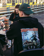 Quattro persone sedute attorno a un tavolo pieghevole all’aperto, mentre giocano a un gioco di impilamento di blocchi. Sullo sfondo, dietro una ringhiera metallica, si vede un treno giallo e bianco. La persona in primo piano indossa la Jägermeister Tradition Hoodie e un Jägermeister Dad Cap verde.