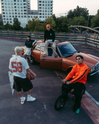 Un gruppo di quattro persone in un contesto urbano all’aperto con un’auto arancione arrugginita, moderni grattacieli e un ponte sullo sfondo. Una persona con una felpa arancione brillante è seduta su una bicicletta in primo piano, un’altra con una maglia bianca Jägermeister con '56' tiene in mano un pallone da calcio, e altre due persone sono posizionate attorno all’auto d’epoca.