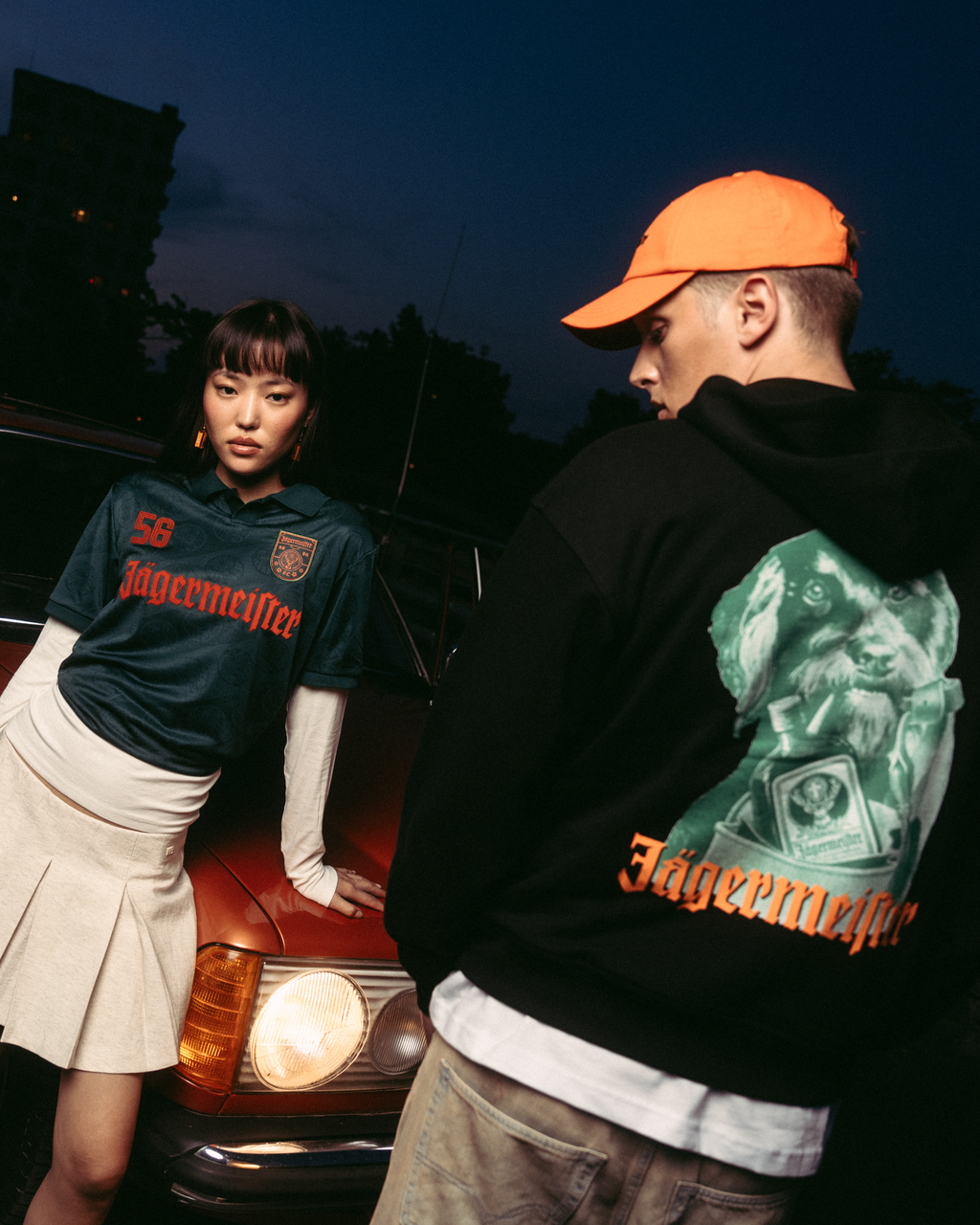 Due modelle in posa davanti a un’auto rossa vintage al crepuscolo. Una modella indossa una jersey Jägermeister verde scuro con una gonna bianca plissettata, e l’altra indossa una felpa con cappuccio nera Jägermeister Waldi e un cappellino Jägermeister Dad arancione.