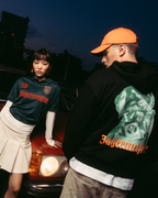 Due modelle in posa davanti a un’auto rossa vintage al crepuscolo. Una modella indossa una jersey Jägermeister verde scuro con una gonna bianca plissettata, e l’altra indossa una felpa con cappuccio nera Jägermeister Waldi e un cappellino Jägermeister Dad arancione.