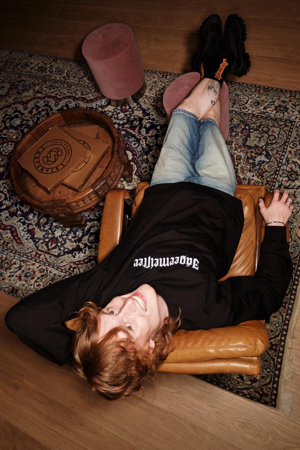 Persona sdraiata su una poltrona in pelle con i piedi sollevati su un tappeto, indossando un crewneck nero Jägermeister.