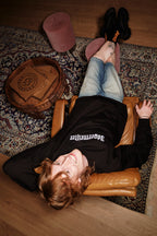 Persona sdraiata su una poltrona in pelle con i piedi sollevati su un tappeto, indossando un crewneck nero Jägermeister.