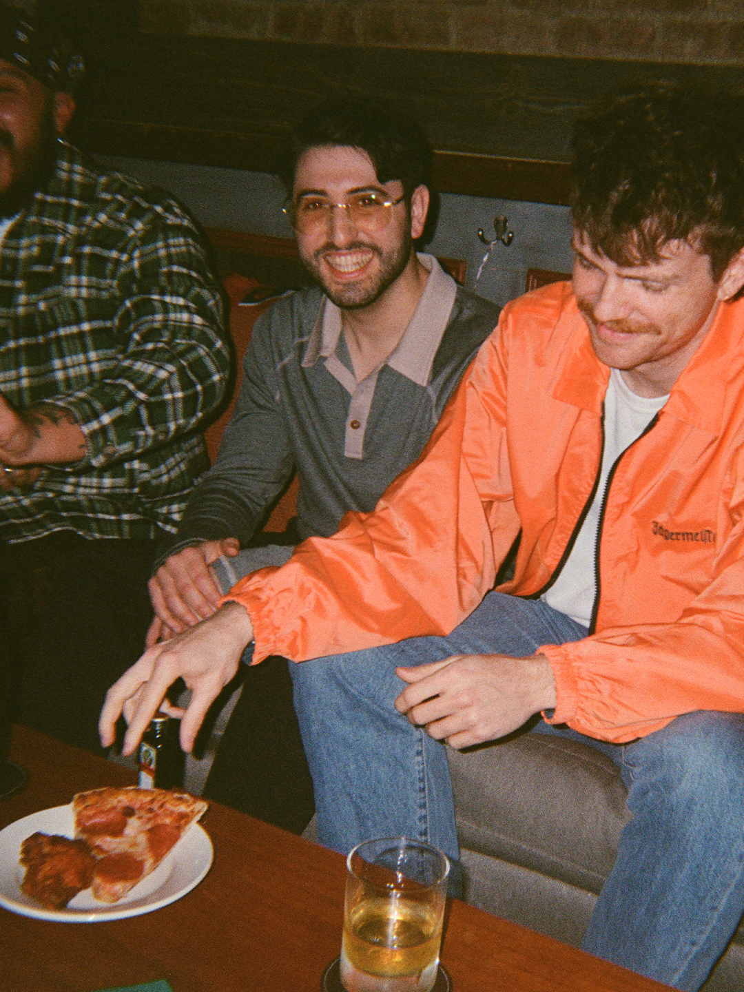 Due uomini seduti insieme, uno indossa una giacca arancione Jägermeister, con un piatto di pizza e un bicchiere su un tavolo.