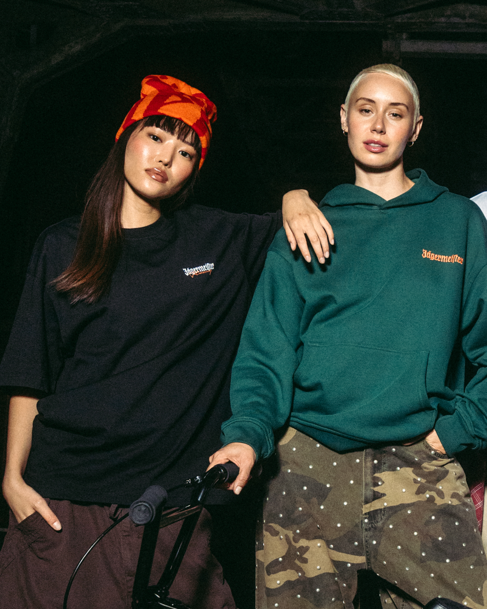 Due giovani donne in posa con una bici BMX nera davanti a un ponte industriale scuro di notte. Sono vestite con capi streetwear Jägermeister, tra cui una felpa verde, una t-shirt bianca, un gilet nero e pantaloni cargo.