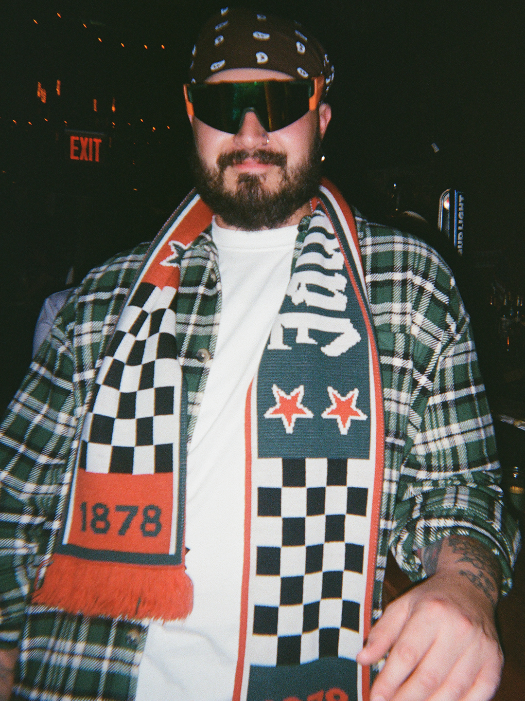 Persona con giacca a quadri, sciarpa Jägermeister Racing, occhiali da sole e cappellino.