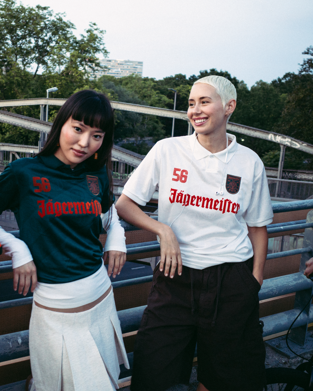 Quattro persone in piedi all’aperto su un ponte o passaggio pedonale, vestite in stile streetwear. Una persona indossa una giacca arancione brillante con zip, un’altra una jersey Jägermeister verde scuro con una gonna bianca, una terza una jersey Jägermeister bianca con pantaloncini scuri, e una quarta una felpa con cappuccio nera e un berretto arancione.