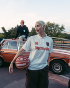 Un gruppo di quattro persone posa all’aperto su un ponte o cavalcavia con un’auto d’epoca arancione dietro di loro. La figura centrale tiene in mano un pallone da basket e indossa una jersey Jägermeister bianca, mentre le altre indossano streetwear arancione brillante, verde e nero.