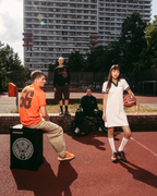 Scena lifestyle streetwear all’aperto su un campo da basket con un alto palazzo sullo sfondo. Un uomo seduto con una jersey Jägermeister arancione è seduto su un box nero tipo speaker, mentre una donna in piedi con un abito polo Jägermeister bianco tiene in mano un pallone da basket.