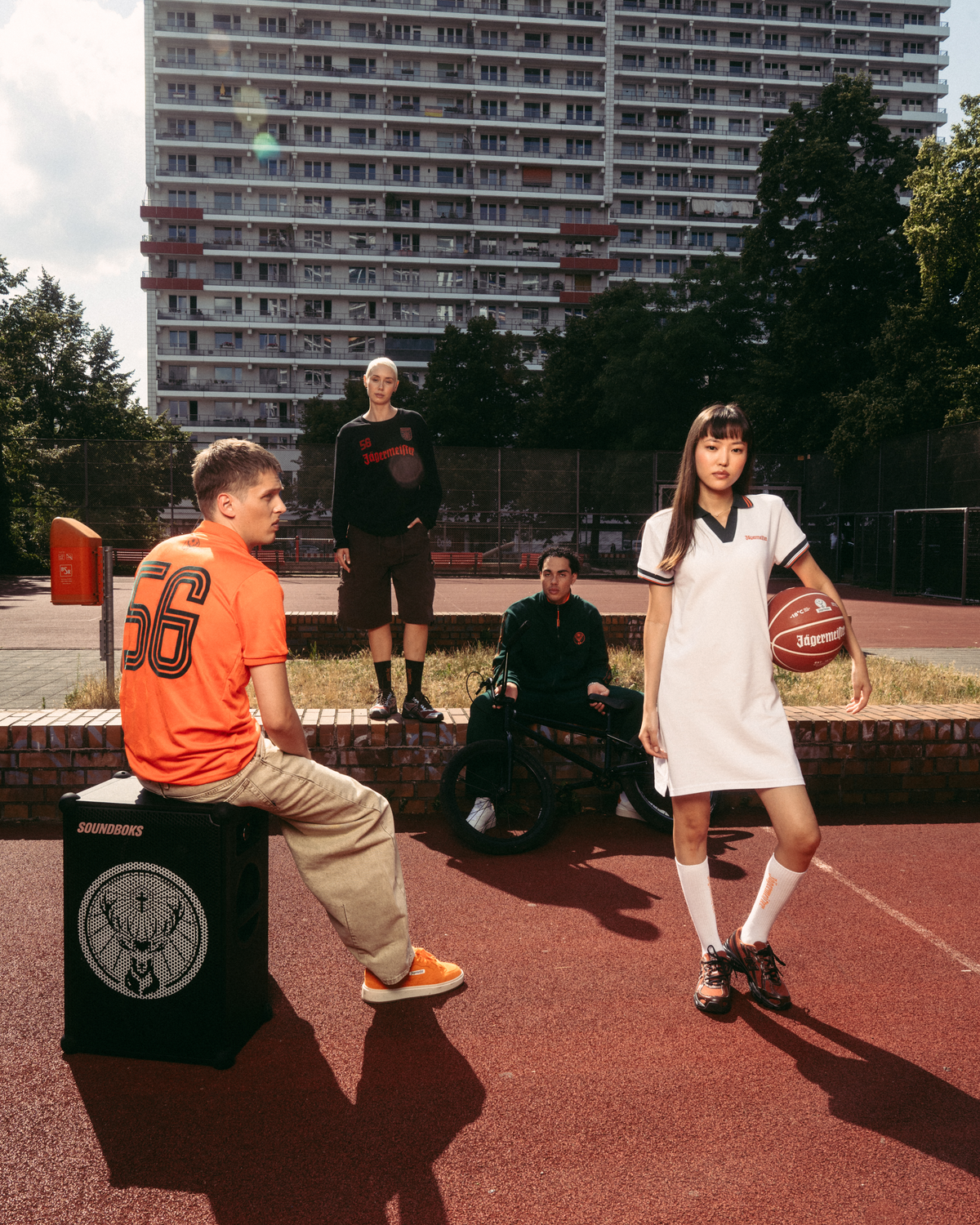 Scena lifestyle streetwear all’aperto su un campo da basket con un alto palazzo sullo sfondo. Un uomo seduto con una jersey Jägermeister arancione è seduto su un box nero tipo speaker, mentre una donna in piedi con un abito polo Jägermeister bianco tiene in mano un pallone da basket.