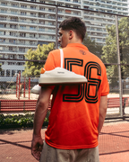 Una persona è in piedi su un campo sportivo all’aperto con una jersey Jägermeister arancione brillante con un grande “56” stampato sul retro. Una paio di sneakers Jägermeister bianche è appoggiato su una spalla, con una recinzione a rete metallica e palazzi sullo sfondo.