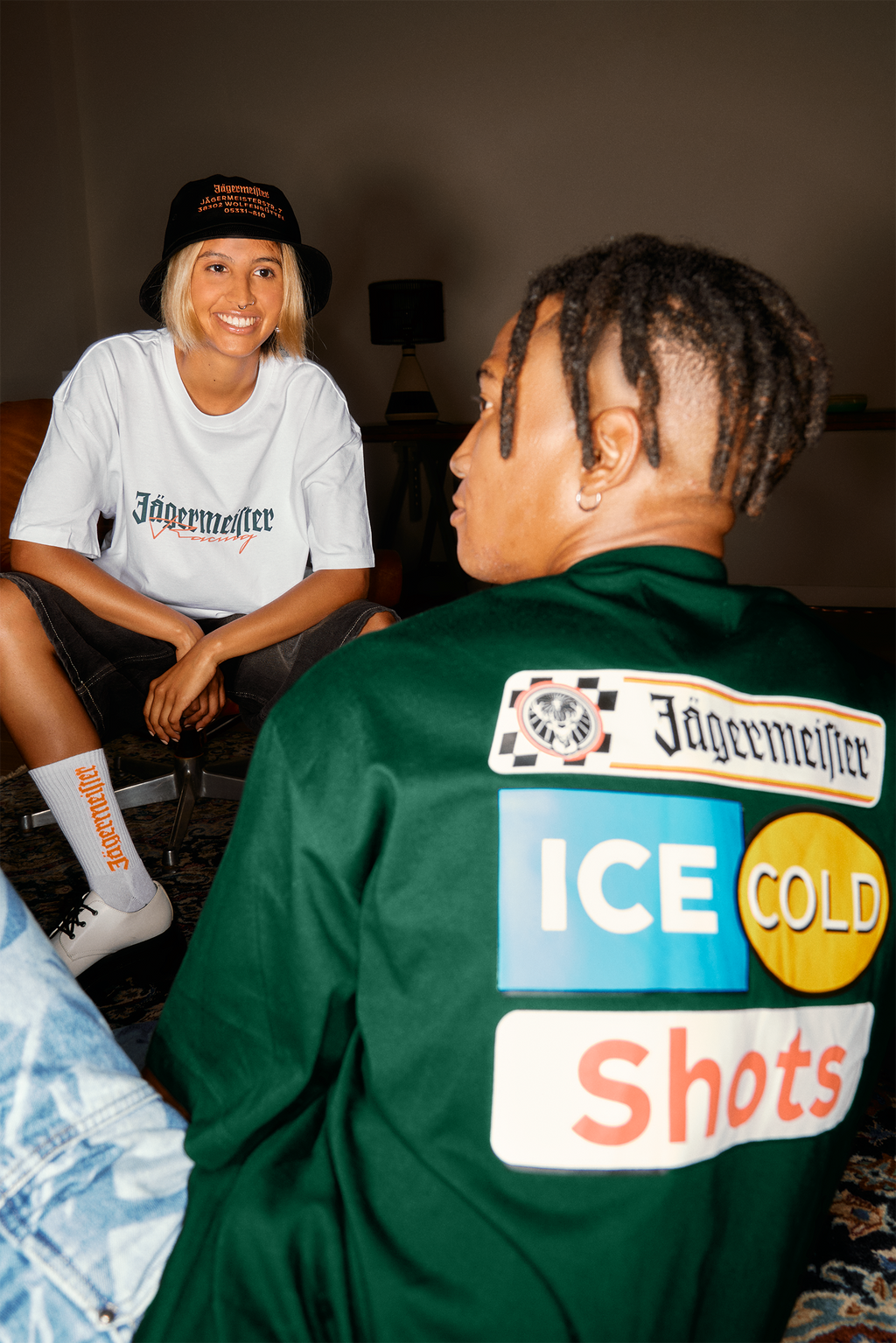 Due persone sedute in casa, una indossa una camicia bianca con branding Jägermeister, l’altra una giacca verde con Jägermeister.