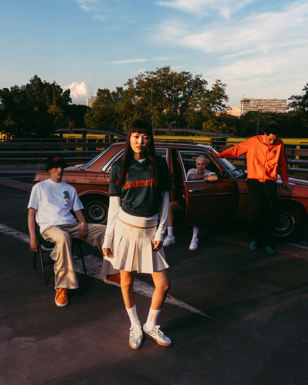 Un gruppo di quattro giovani posa all’aperto su un circuito al tramonto accanto a un’auto d’epoca rossa. La figura centrale indossa una jersey Jäermeister verde scuro con scritte arancioni, una gonna bianca plissettata e sneakers bianche.