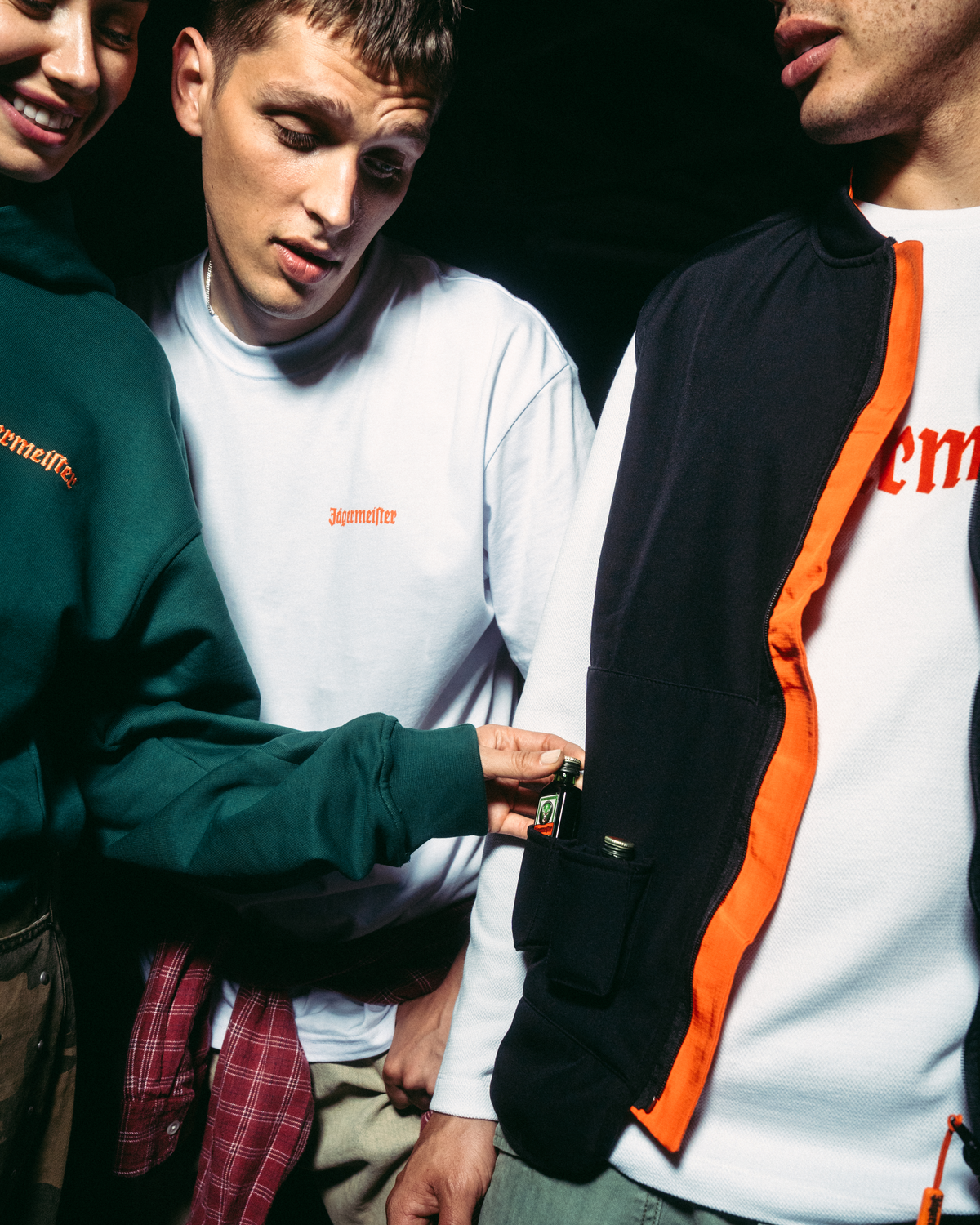 Tre giovani in piedi vicini in un ambiente scuro, con capi Jägermeister. Uno indossa una felpa con cappuccio verde scuro, un altro una maglia bianca a maniche lunghe e il terzo una giacca nera e arancione con zip sopra una maglia bianca.