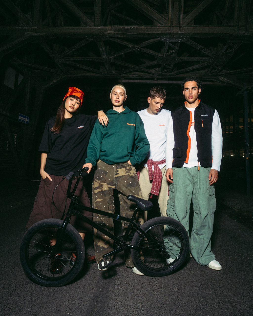 Quattro giovani in posa con una bici BMX nera in un ambiente notturno, scuro e industriale sotto un ponte. Indossano outfit streetwear Jägermeister, tra cui una felpa con cappuccio verde, una T-shirt bianca, un gilet nero e pantaloni cargo.