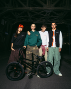 Quattro giovani in posa con una bici BMX nera in un ambiente notturno, scuro e industriale sotto un ponte. Indossano outfit streetwear Jägermeister, tra cui una felpa con cappuccio verde, una T-shirt bianca, un gilet nero e pantaloni cargo.