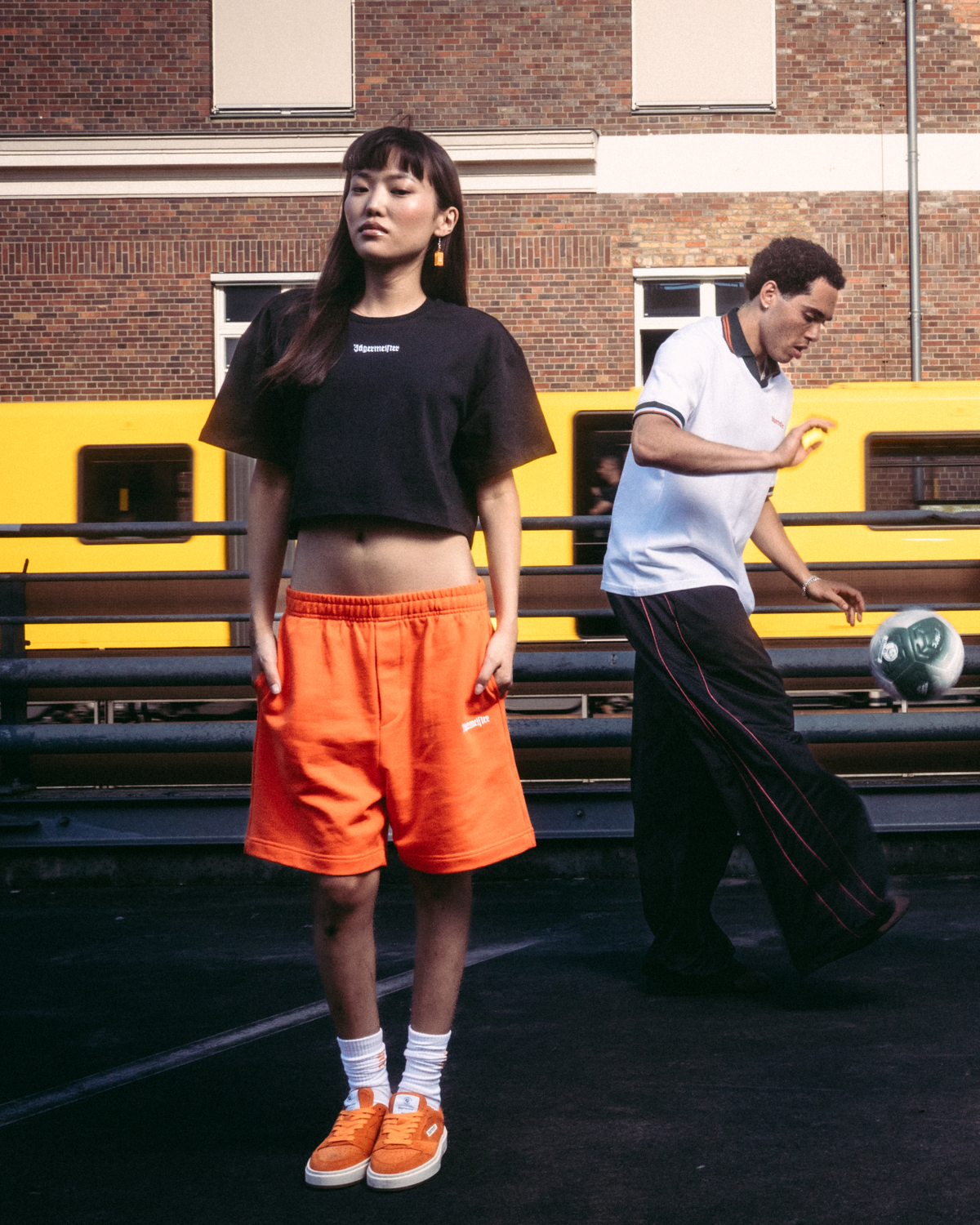 Quattro modelli che indossano capi streetwear Jägermeister, tra cui shorts arancioni, un crop top nero, una t-shirt bianca e una felpa con cappuccio nera con stampa grafica sul retro, in piedi su una superficie asfaltata scura davanti a un edificio in mattoni e a un treno giallo.