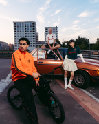 Tre modelle in outfit streetwear Jägermeister in posa con una vintage car arancione e una BMX nera su un tetto urbano al tramonto. Gli outfit includono una giacca zip-up Jägermeister arancione acceso, pantaloni da tuta neri con strisce rosse, una jersey bianca Jägermeister, una jersey verde Jägermeister con branding arancione e una gonna bianca plissettata.