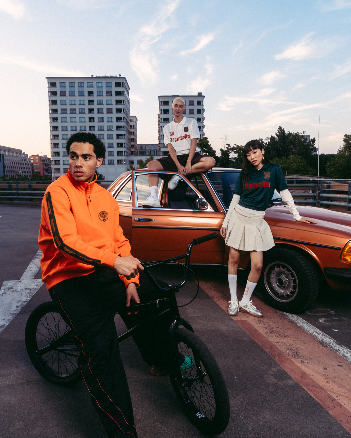 Tre modelle in outfit streetwear Jägermeister in posa con una vintage car arancione e una BMX nera su un tetto urbano al tramonto. Gli outfit includono una giacca zip-up Jägermeister arancione acceso, pantaloni da tuta neri con strisce rosse, una jersey bianca Jägermeister, una jersey verde Jägermeister con branding arancione e una gonna bianca plissettata.