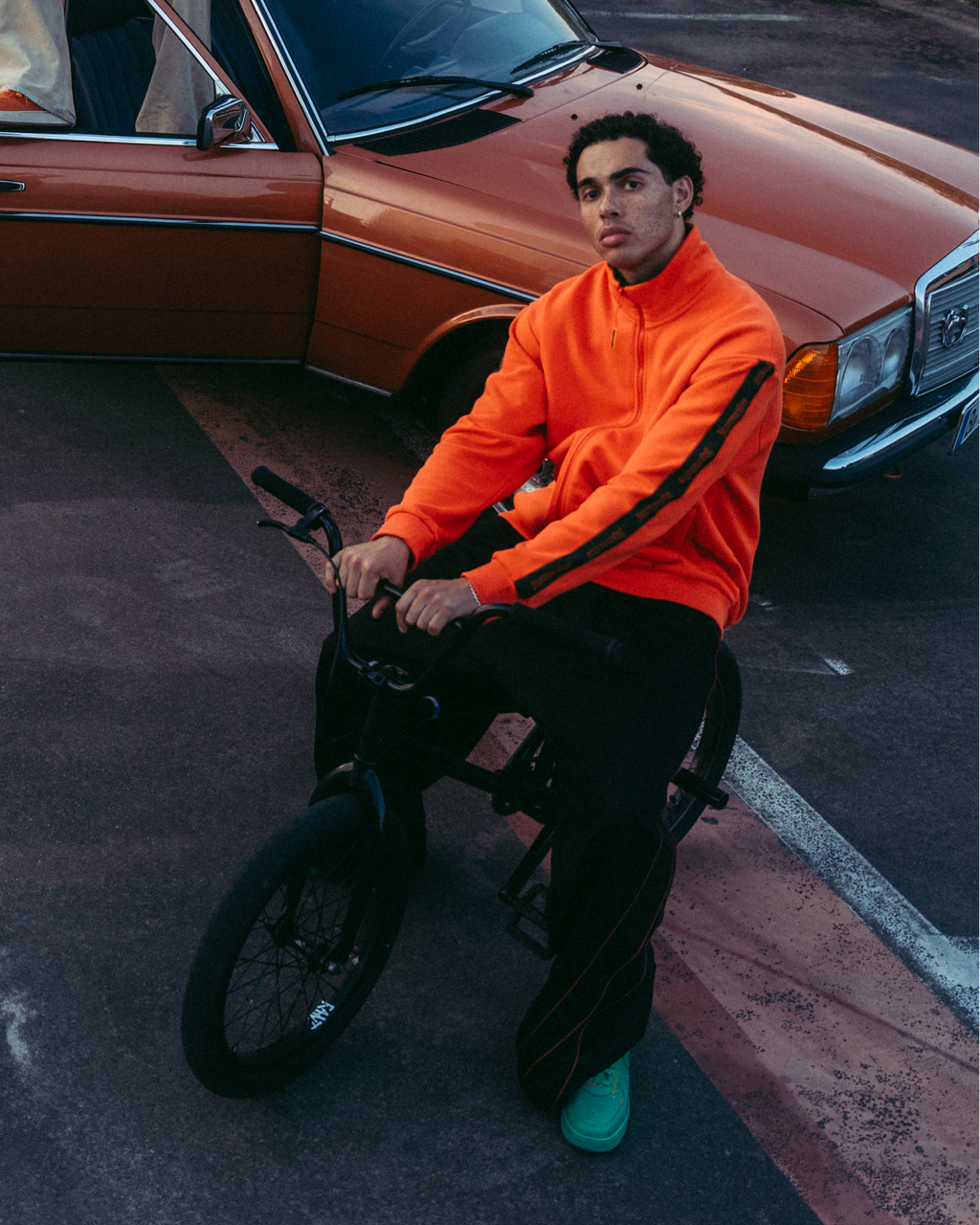 Un gruppo di quattro persone in un ambiente urbano all’aperto con un’auto arancione arrugginita, grattacieli moderni e un ponte sullo sfondo. Una persona con una zip-up Jägermeister arancione acceso è seduta su una bici in primo piano.