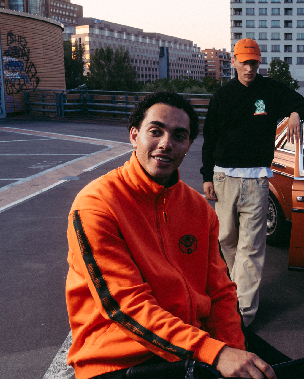 Quattro modelle in posa su un tetto urbano con una vintage car arancione, indossando vari outfit streetwear Jägermeister, tra cui una zip-up Jägermeister arancione acceso, una T-shirt bianca Jägermeister con Waldi, una jersey Jägermeister verde scuro e una gonna bianca plissettata.