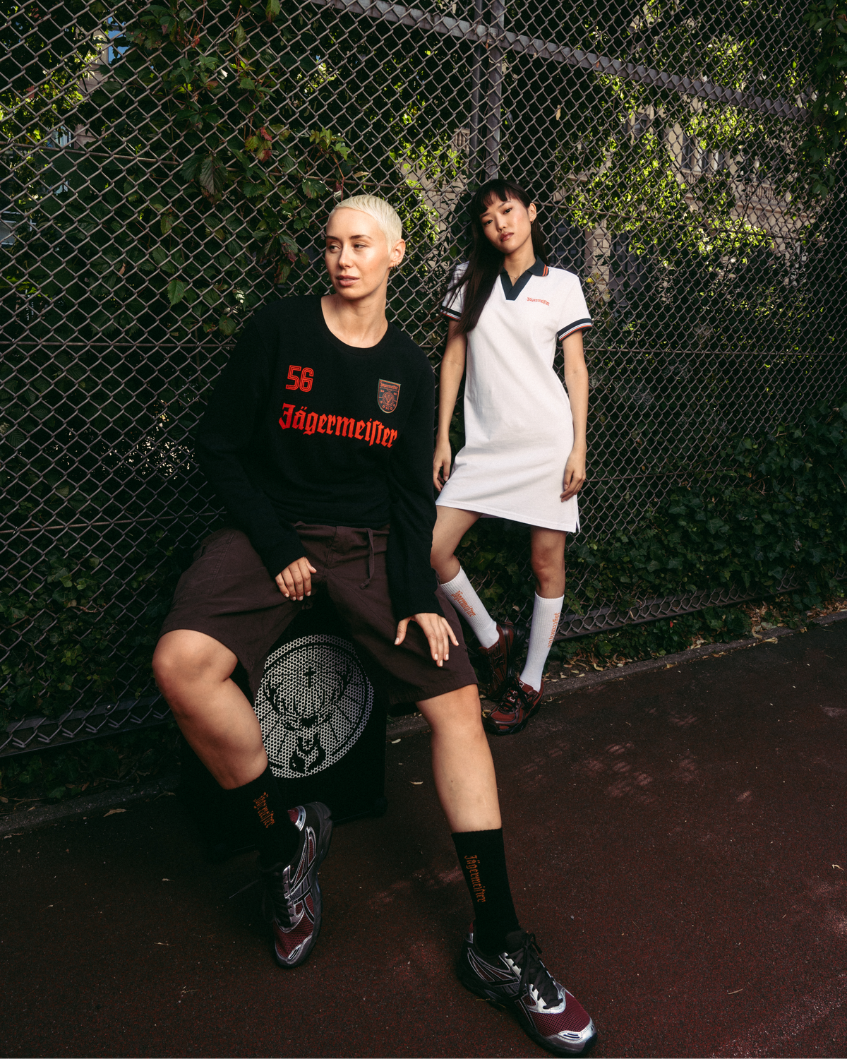 Due modelle posano davanti a una recinzione a maglie metalliche. La modella a sinistra indossa una maglia nera a maniche lunghe Jägermeister, pantaloncini marroni e calze nere.