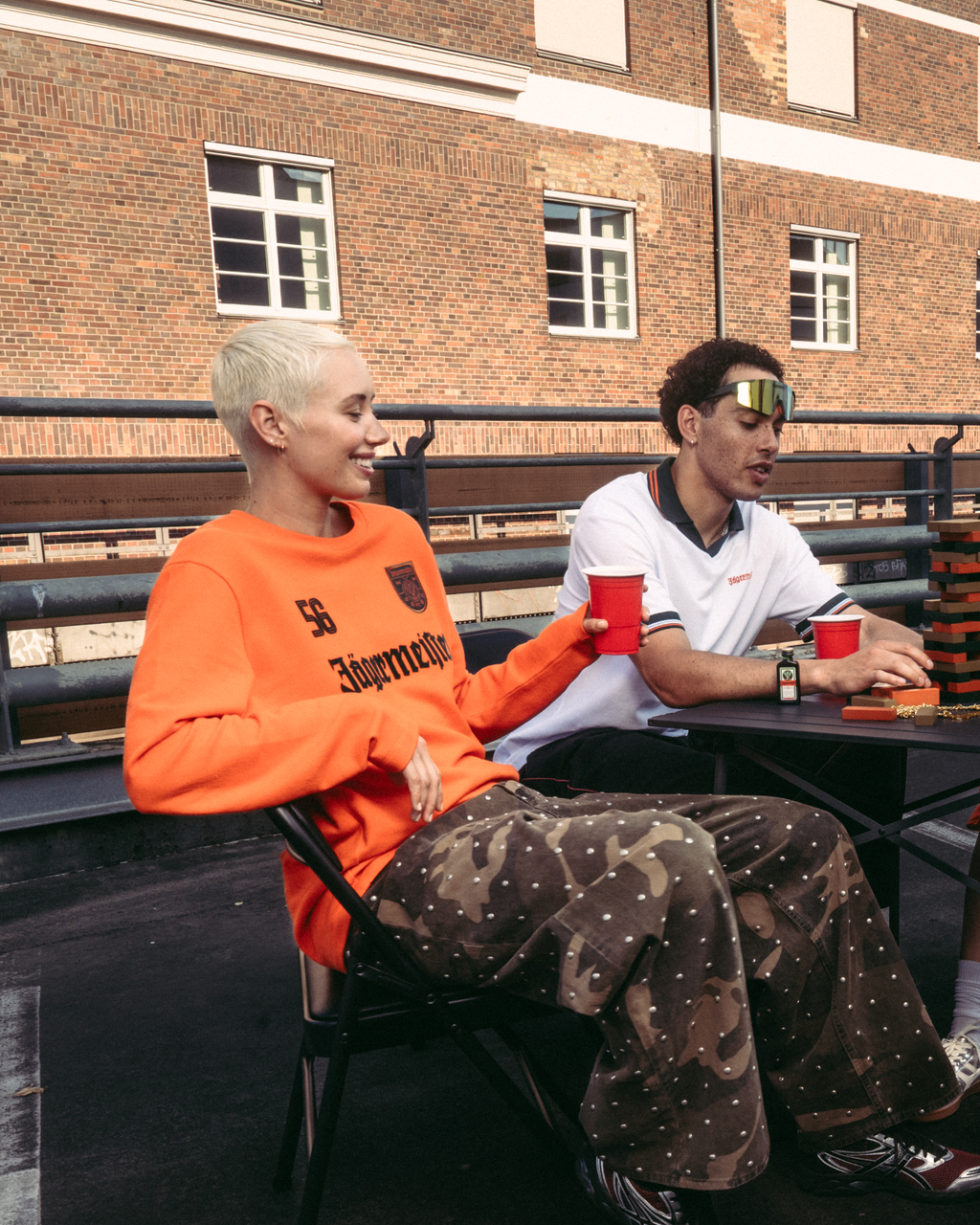 Quattro persone sedute attorno a un piccolo tavolo su un rooftop all’aperto o su un ponte, con bicchieri rossi in mano. Sono vestite con casual streetwear Jägermeister, tra cui una maglia a maniche lunghe arancione e una polo bianca.