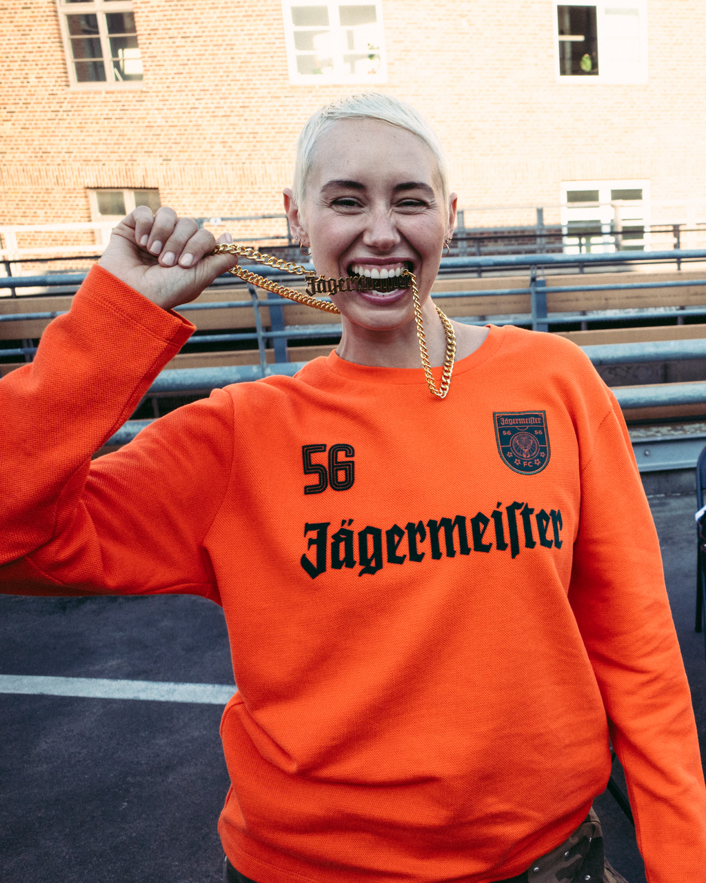 In primo piano una donna sorridente indossa una felpa arancione brillante a maniche lunghe Jägermeister con '56 Jägermeister' stampato in nero sul davanti, e tiene in mano una catena dorata Jägermeister. 