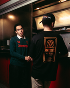 Due uomini in piedi fuori da un food truck rosso di notte. Uno dei due guarda la camera e indossa una maglia a maniche lunghe Jägermeister verde scuro con testo arancione, pantaloni neri e sneakers bianche.