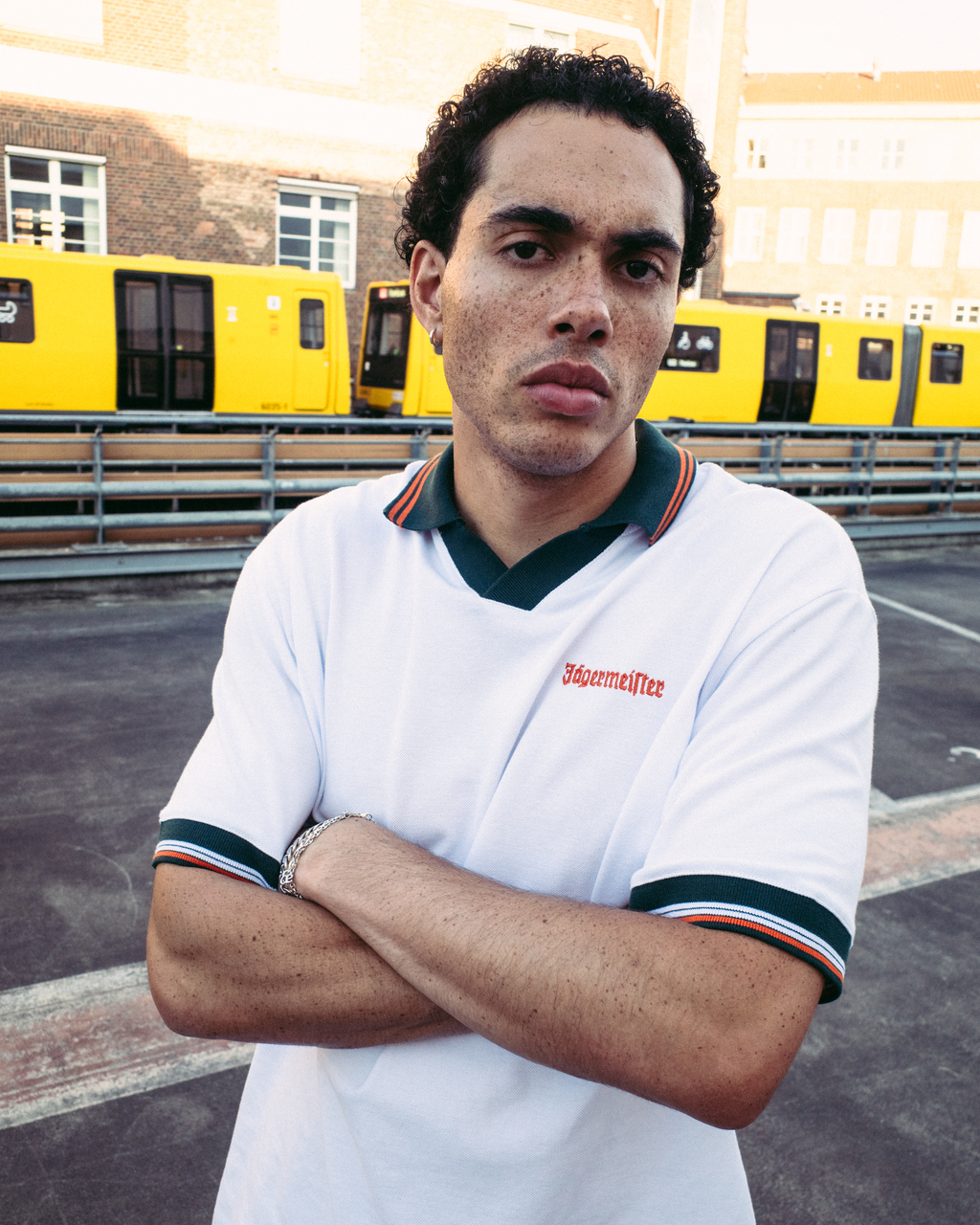 Un uomo con una polo bianca a maniche corte Jägermeister con rifiniture verde scuro e arancioni, con il nome del marchio 'Jägermeister' sul petto, in piedi a braccia incrociate davanti a dei treni gialli e a un edificio in mattoni.