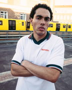 Un uomo con una polo bianca a maniche corte Jägermeister con rifiniture verde scuro e arancioni, con il nome del marchio 'Jägermeister' sul petto, in piedi a braccia incrociate davanti a dei treni gialli e a un edificio in mattoni.