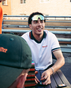 Un uomo con una polo bianca e verde Jägermeister sorride mentre è seduto all’aperto a un tavolo, con un’altra persona in primo piano che indossa un cappellino verde Jägermeier e un edificio in mattoni sullo sfondo.