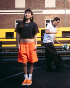 Quattro modelli che indossano capi streetwear Jägermeister, tra cui shorts arancioni, un crop top nero, una t-shirt bianca e una felpa con cappuccio nera con stampa grafica sul retro, in piedi su una superficie asfaltata scura davanti a un edificio in mattoni e a un treno giallo.
