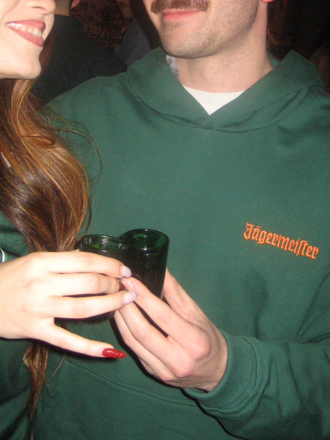 Persona con felpa con cappuccio verde Jägermeister e wordmark Jägermeister arancione sul petto.