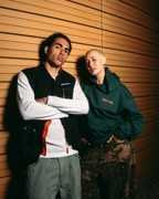 Due modelle in posa davanti a una parete rivestita in legno caldo. La modella a sinistra indossa un gilet nero Jägermeister sopra una camicia bianca a maniche lunghe con dettagli arancioni e pantaloni cargo verde oliva.