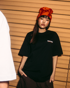 Due modelli in posa davanti a una parete in listelli di legno. La modella a sinistra mostra il retro di una t-shirt bianca con una grande stampa grafica arancione che recita 'Jägermeister'.