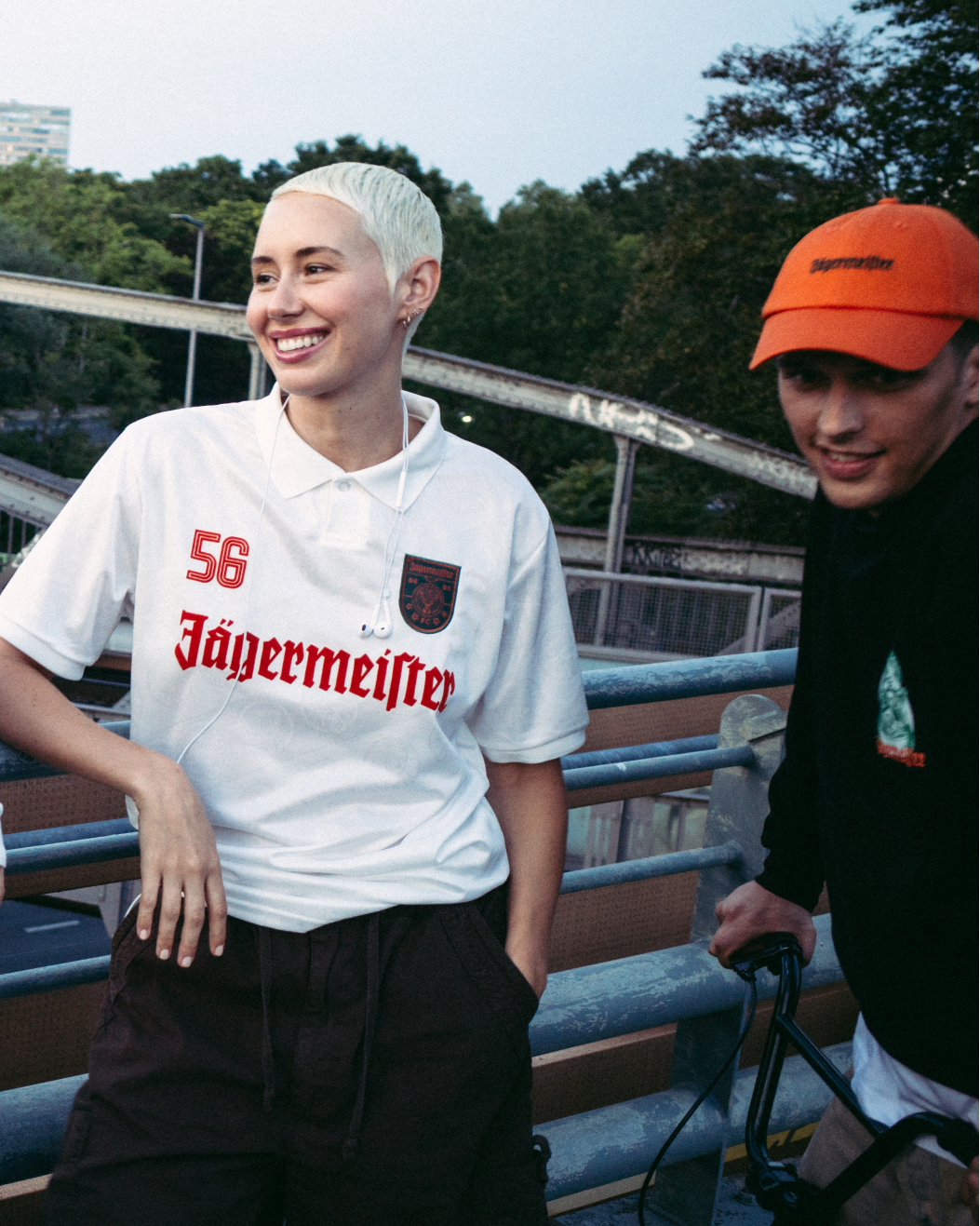 Quattro persone in piedi all'aperto su un ponte o una passerella, con streetwear Jägermeister. Una indossa una giacca arancione brillante con zip, un'altra una maglia verde scuro Jagermeister con una gonna bianca, una terza una maglia bianca Jagermeister con pantaloncini scuri e la quarta una felpa nera Jägermeister e un berretto arancione.
