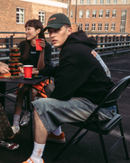 Quattro persone sedute attorno a un piccolo tavolo su un rooftop o un ponte all'aperto, con in mano bicchieri rossi. Indossano streetwear Jägermeister casual, tra cui una felpa arancione, una t-shirt bianca, una felpa con cappuccio nera e pantaloni fantasia.