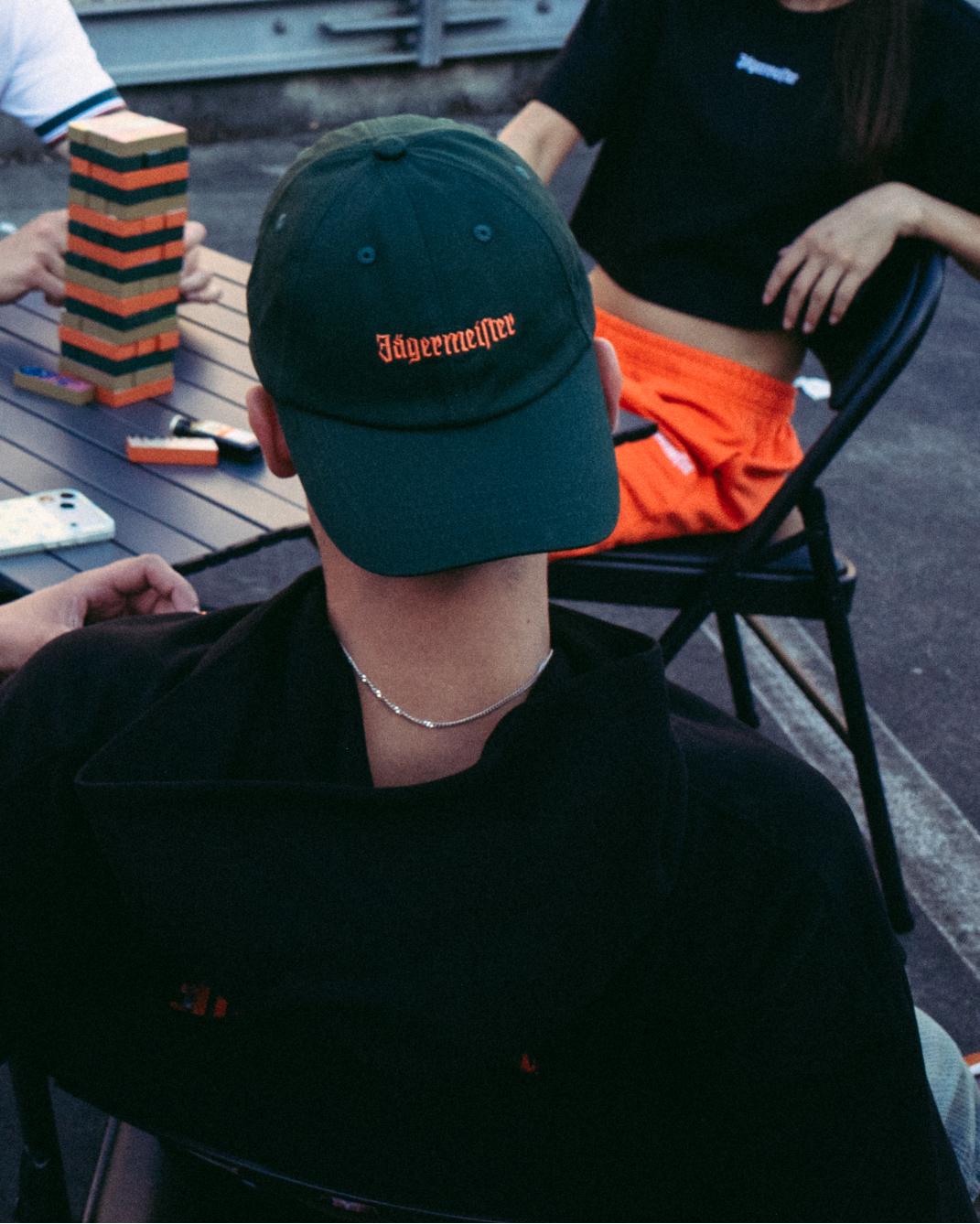 Una donna sorridente in primo piano indossa una felpa arancione brillante a maniche lunghe con '56 Jägermeister' stampato in nero sul davanti e tiene in mano una catena d'oro. Sullo sfondo, tre persone sono sedute a un tavolo su un rooftop o una terrazza all'aperto, con un edificio in mattoni dietro di loro.