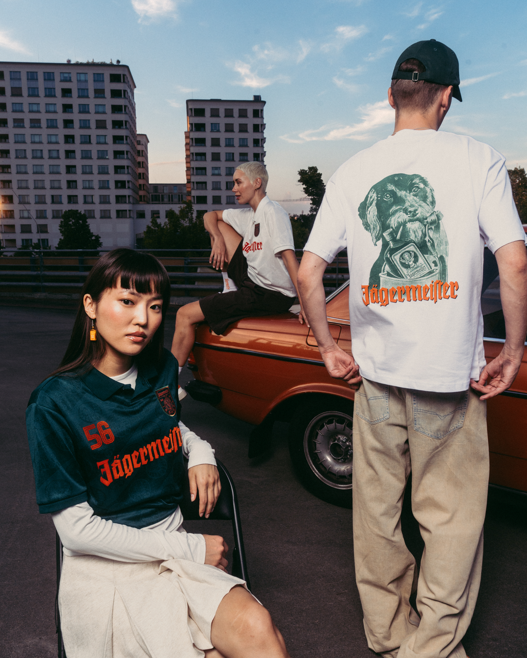 Tre modelle e modelli indossano capi Jägermeister in un contesto urbano all’aperto, con un’auto d’epoca arancione e palazzi sullo sfondo. Una modella è seduta in primo piano con una jersey Jägermeister verde scuro, un altro è in piedi di spalle con una T-shirt bianca grafica e pantaloni beige, e un terzo modello è seduto sull’auto con una T-shirt bianca.