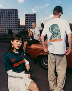 Tre modelle e modelli indossano capi Jägermeister in un contesto urbano all’aperto, con un’auto d’epoca arancione e palazzi sullo sfondo. Una modella è seduta in primo piano con una jersey Jägermeister verde scuro, un altro è in piedi di spalle con una T-shirt bianca grafica e pantaloni beige, e un terzo modello è seduto sull’auto con una T-shirt bianca.