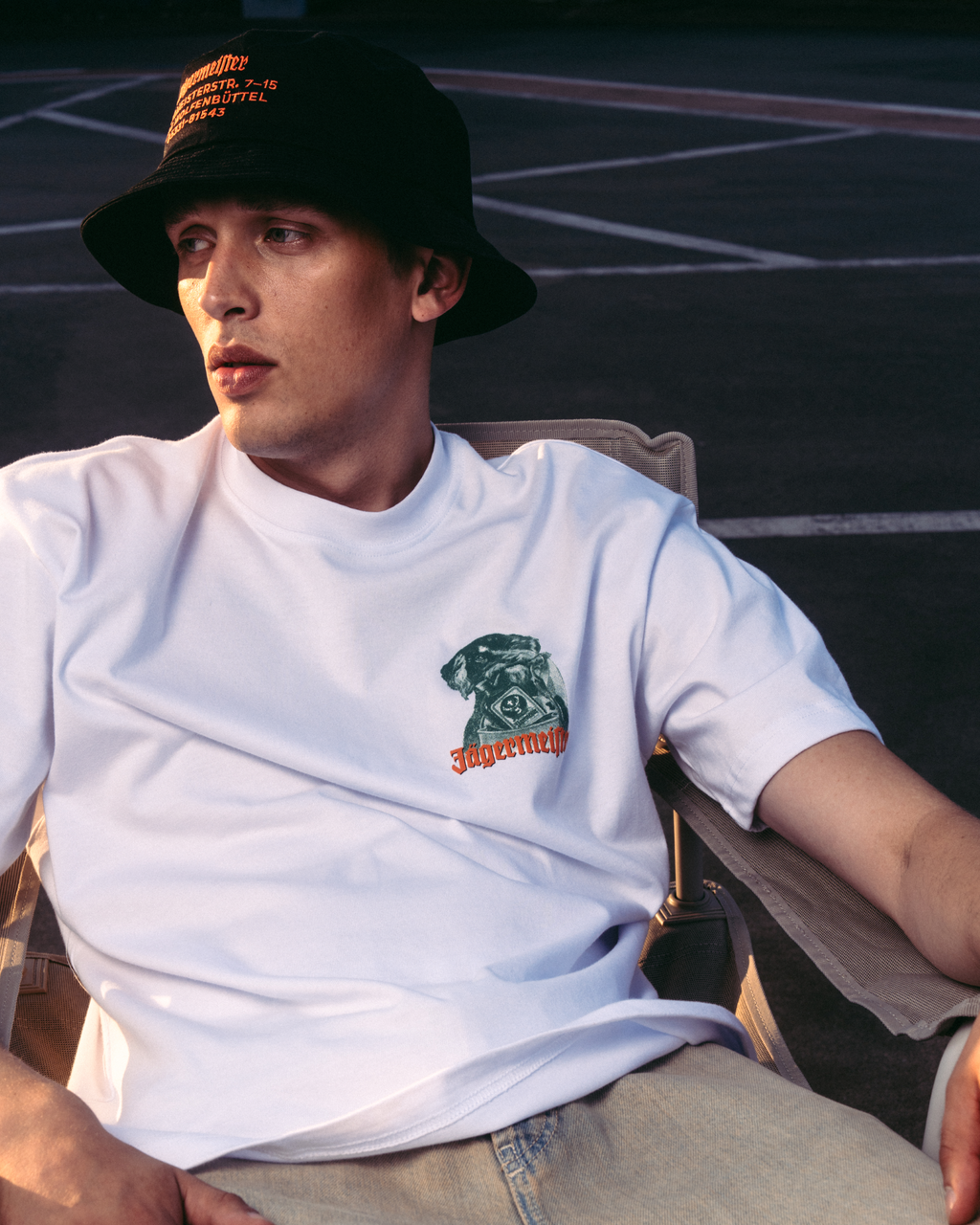 Un uomo con una t-shirt bianca grafica con logo Jägermeister Waldi, un bucket hat nero e jeans beige, seduto su una sedia in un ambiente all’aperto.