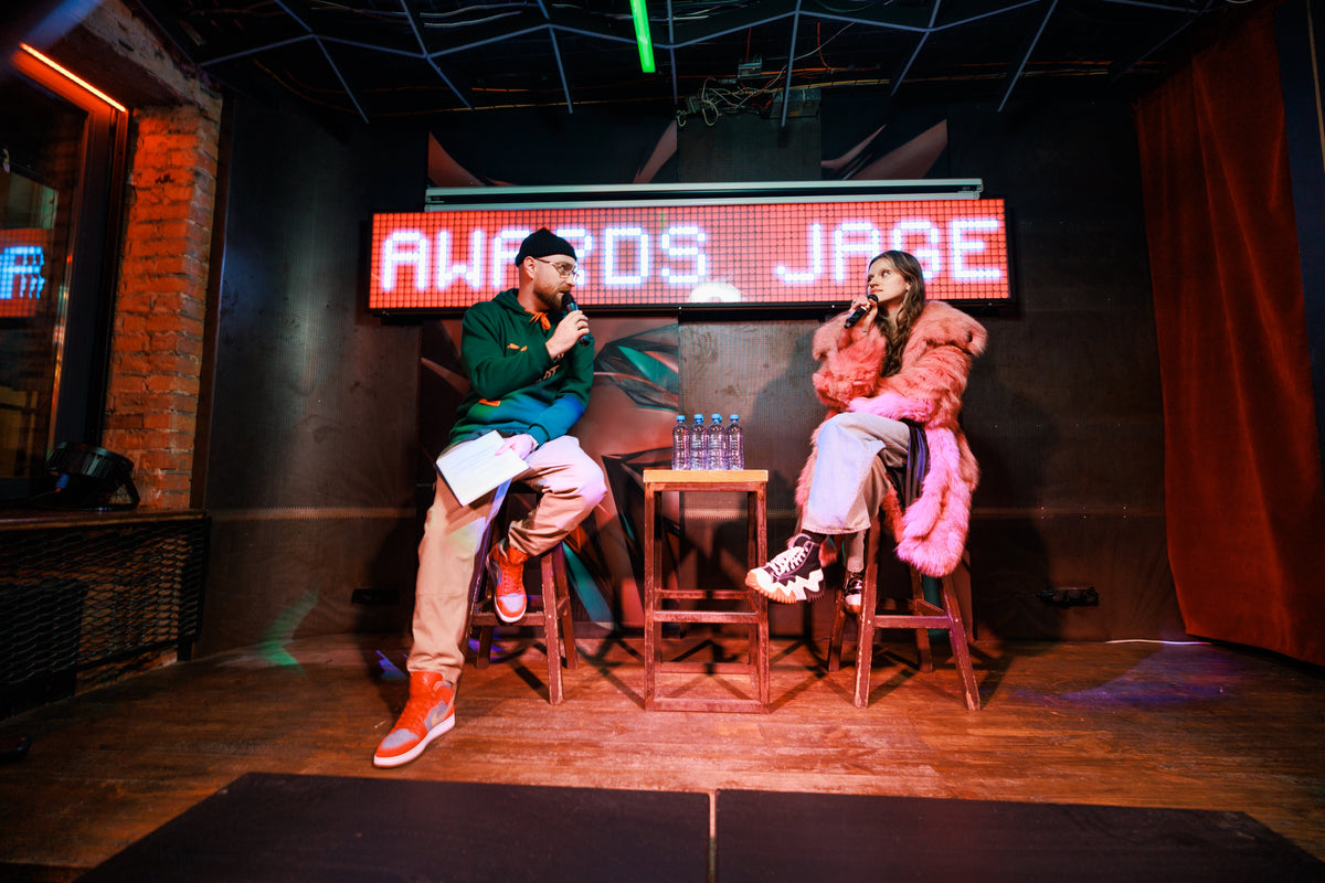 A man and a woman sit on stools on a dimly lit stage, with a large red LED sign behind them reading 'AWARDS JAGE'. The man wears a green jacket, beige pants, and orange sneakers, while the woman wears a pink jacket and light-colored pants.